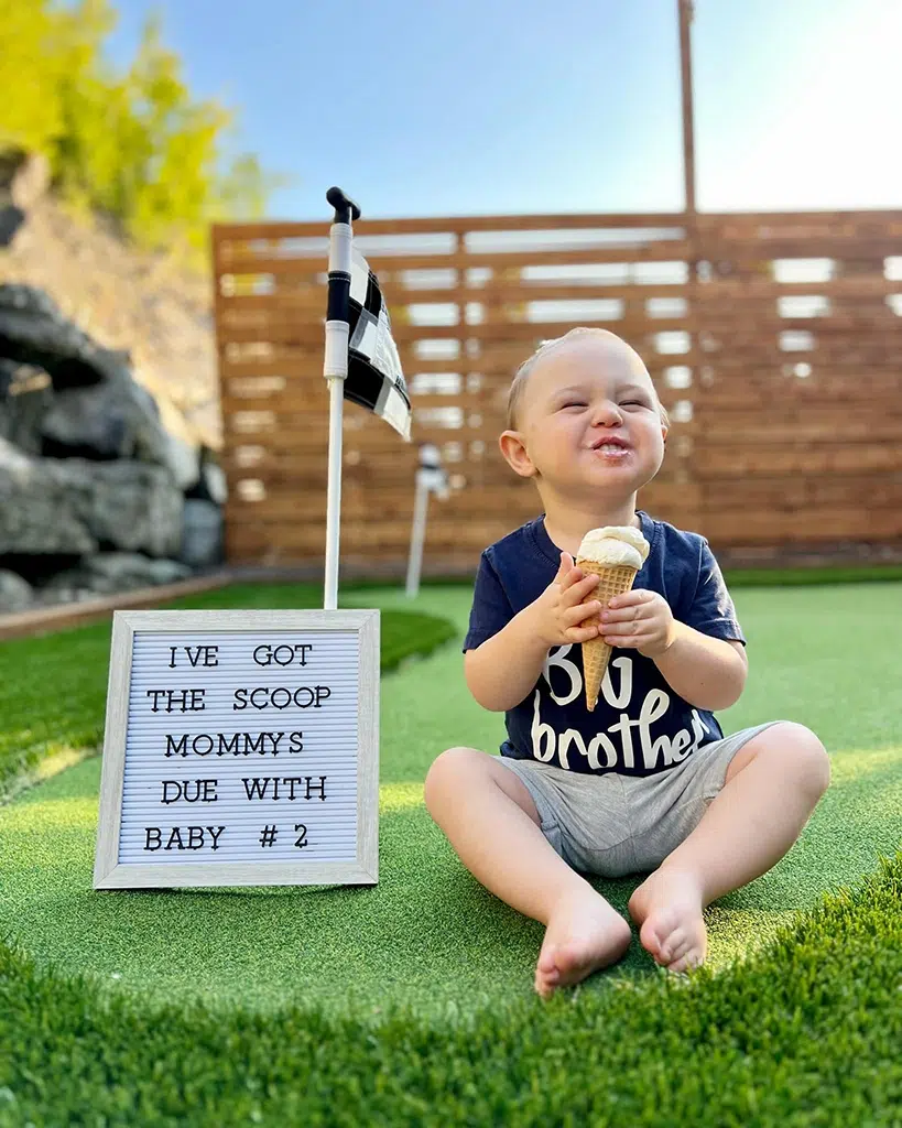 A baby sitting on a green grass with an ice cream sign.